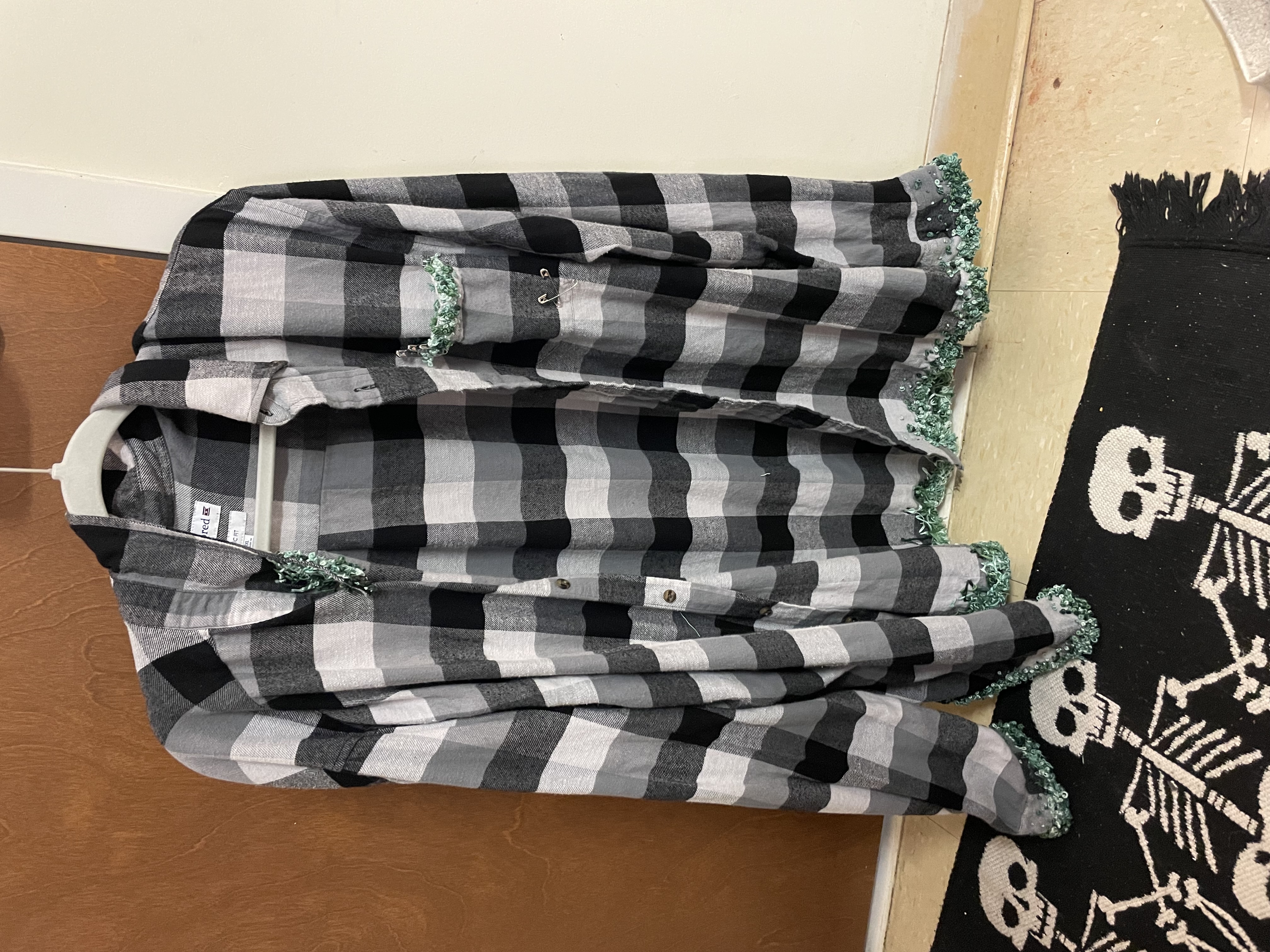 a picture of a black and white flannel shirt with green embrodiery on the cuffs, part of a collar, pocket, and bottom edge. On the chest, there is a pocket. There are saftey pins in the top left of the pocket and two forming an 